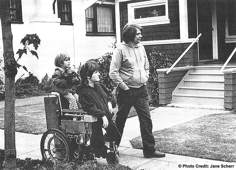 Family of three strolling in the neighborhood. The father is walking. The mother is using a wheelchair and the child is riding on the back of his mothers chair. Photo: Jane Sherr