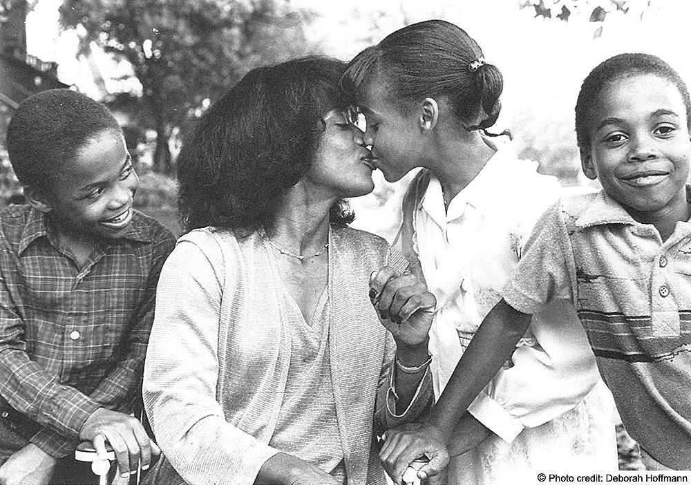 Re’gena with her children. Two boys and a girl. She's giving the girl s kiss. Photo Credit: Deborah Hoffman is embedded in the image.