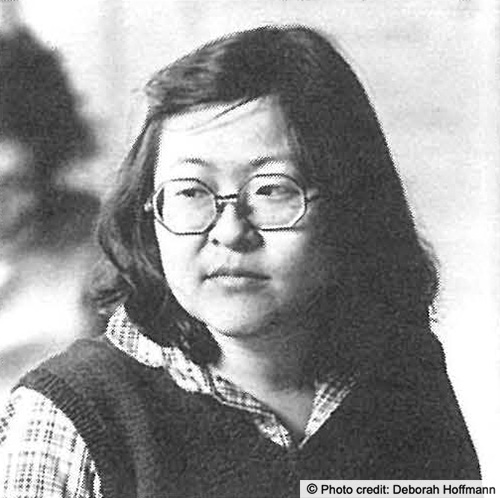 Detail of a photo used earlier of a Woman in motorized wheelchair with notebook in her lap. Here we are focused on her face. She is wearing glasses and has a half smile. Photo credit: Deborah Hoffmann