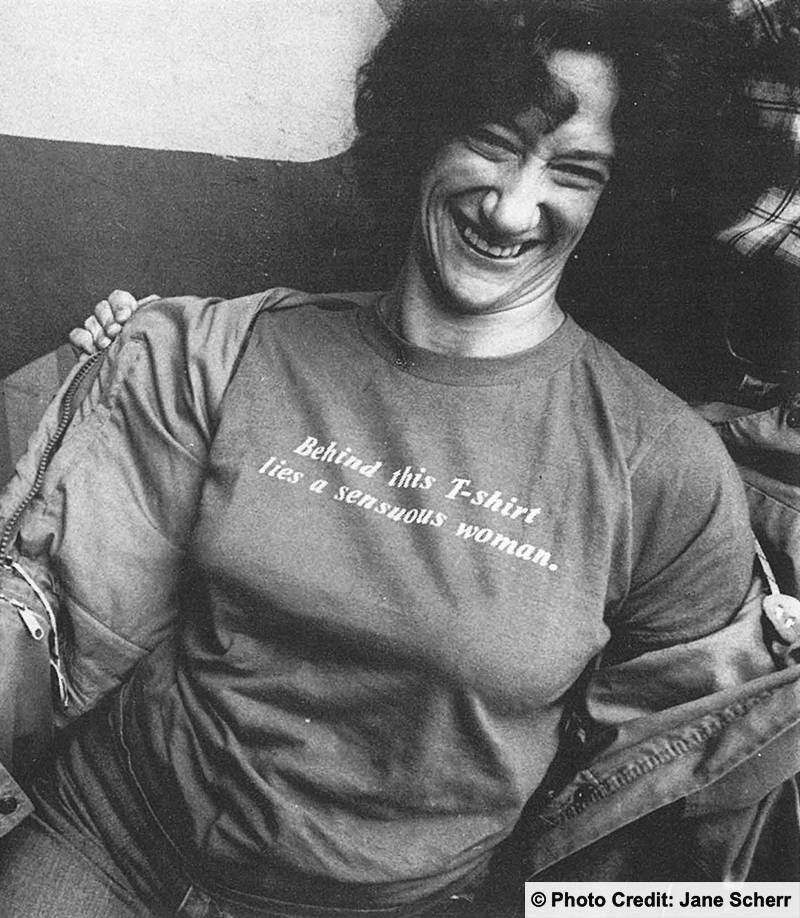 A woman is sitting down laughing taking her jacket off. Her T-shirt reads: Behind this t-shirt is a sensual woman. Photo credit to Jane Scherr