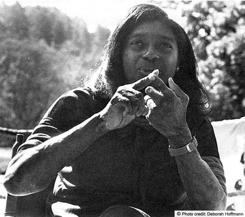 Woman seated outside talking and gesturing with her hands. Her arms appear to have been burned.