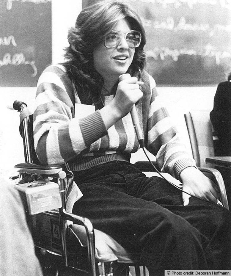 Nancy is seated in a wheelchair and holding a microphone to address an audience. Photo credit to Deborah Hoffman is printed on the image.