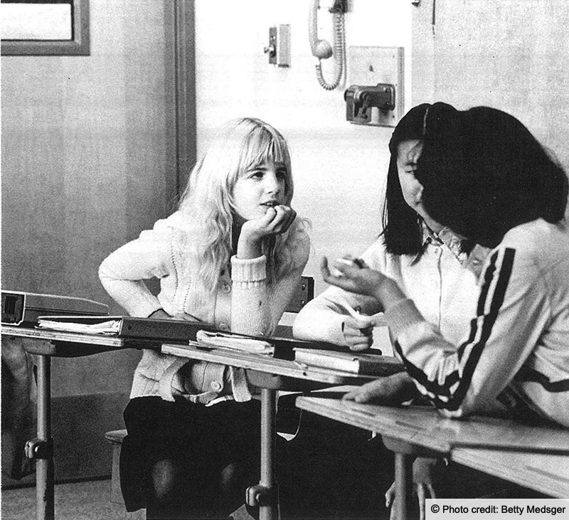 Three young female students chat at their desks in the classroom. Photo credit: Betty Medsger