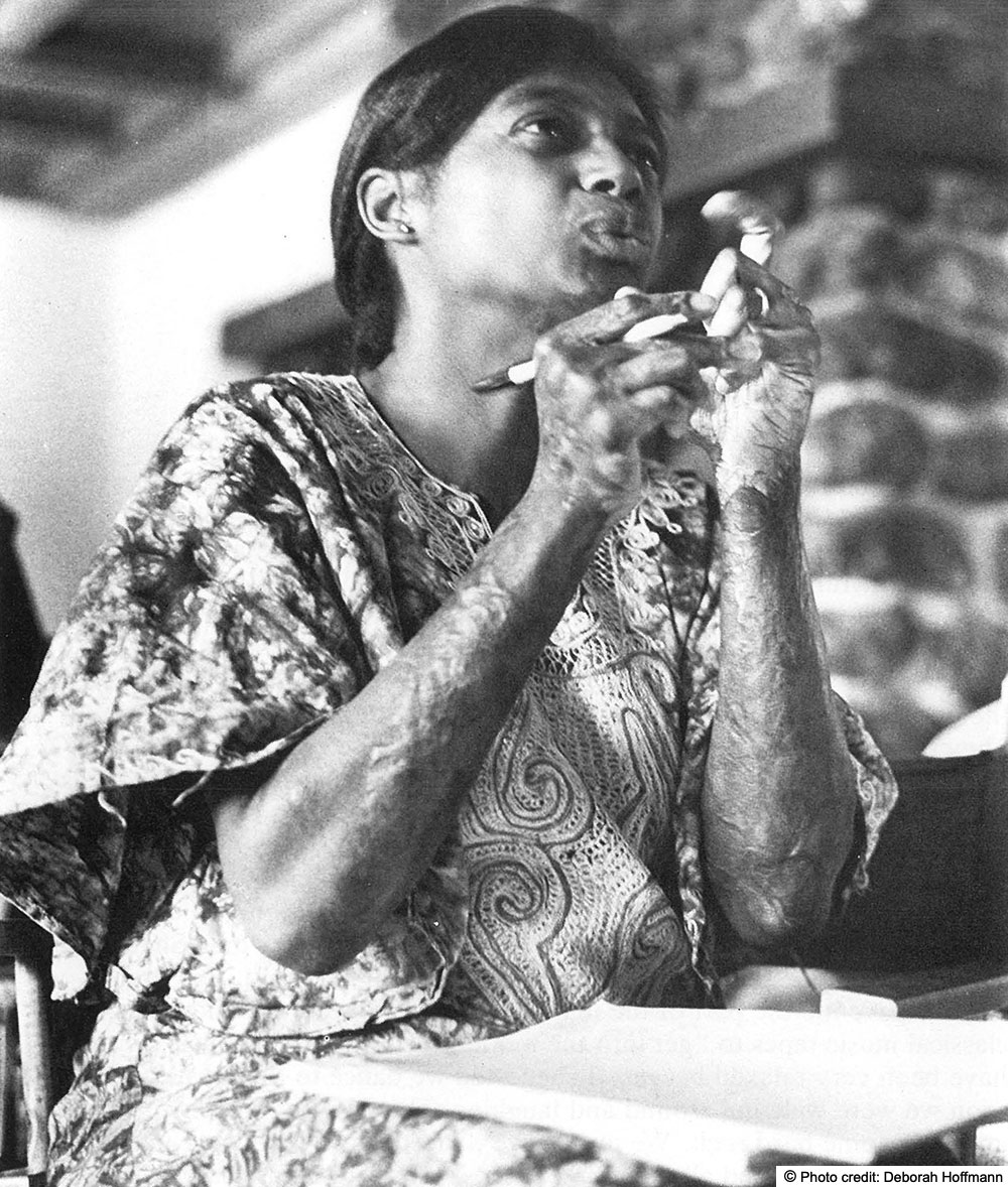 Lois is seated with a notebook in her lap. Her hands are raised, she is holding a pen while she is speaking. Photo Credit: Deborah Hoffman is embedded in the image.