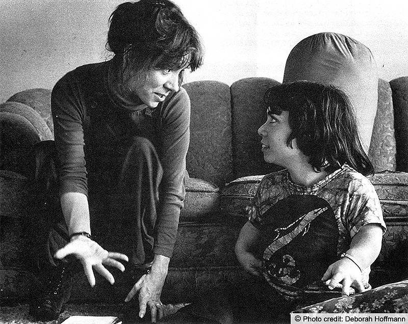 Two women, one a person with dwarfism, sit on the floor in front of a couch talking. Photo Credit: Deborah Hoffman - text is embedded in the image.