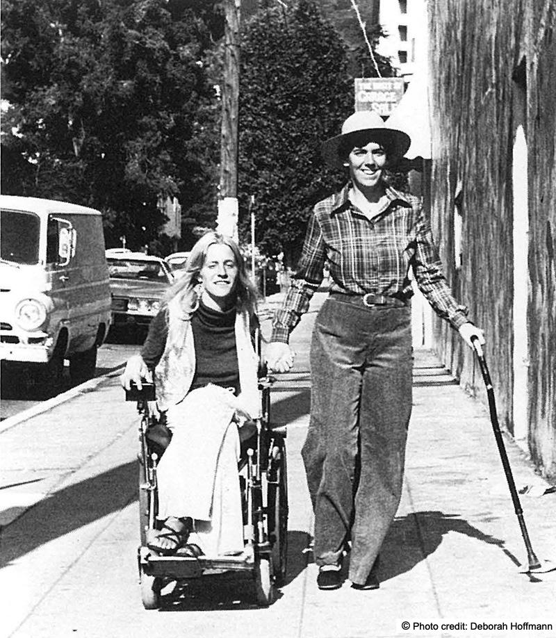 Two young women on a city sidewalk. One is walking using a cane and the other is using a wheelchair. Photo credit: Deborah Hoffman