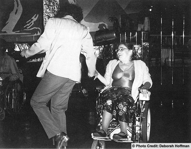 Two people are dancing at a night club. One is a woman and wheelchair user. They are dressed for a night out are having a lot of fun. Photo: Deborah Hoffman