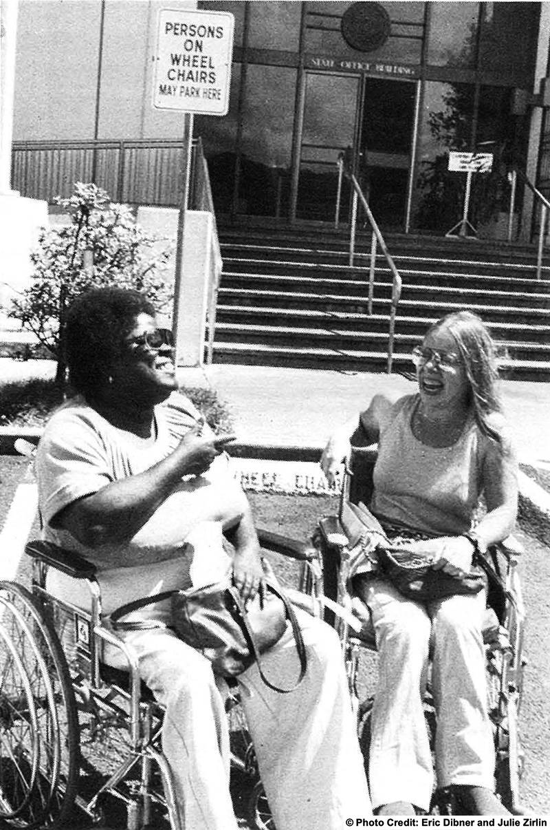 Two wheelchair using woman talk an laugh under a sign that reads: Persons on wheelchairs may park here.