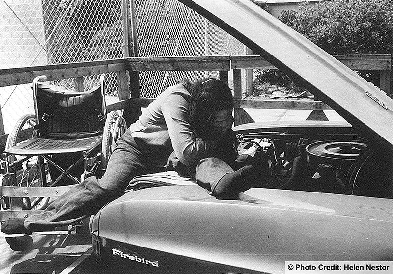 Wheelchair user climbs out of his chair to work on the engine of his car. Photo credit: Helen Nestor