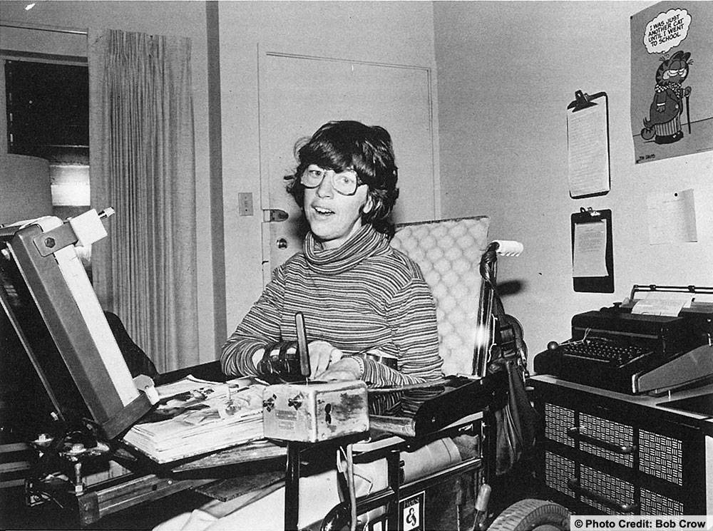 Norma is seated in her wheelchair looking at the camera. A typewriter behind her and clipboards hanging on the wall. Photo Credit: Bob Crow is embedded in the image.