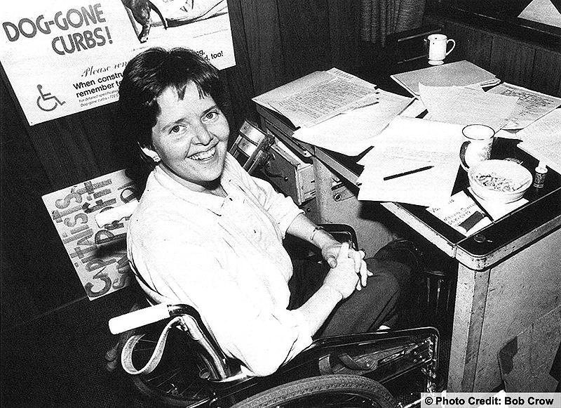 Woman in her wheelchair at her desk smiling at the camera. Desk is covered with papers and coffee cups. Photo credit: Bob Crow