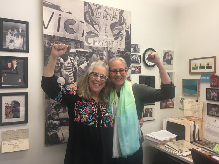 Arlene Mayerson and Larisa Cummings with their arms around each other. Their other arms are raised in the air, their  hands in fists.