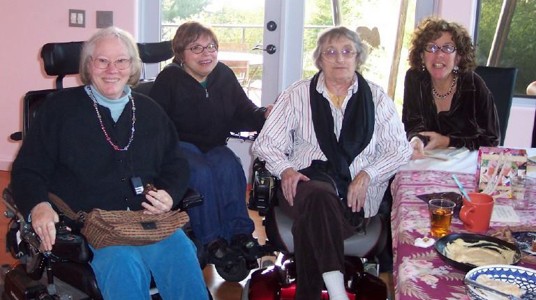 Arlene seated at table. Kitty, Judy and Pam in their wheelchairs next to table. French doors behind them showing a sunny day