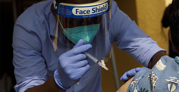 Man with mask and face shield administering a vaccine shot