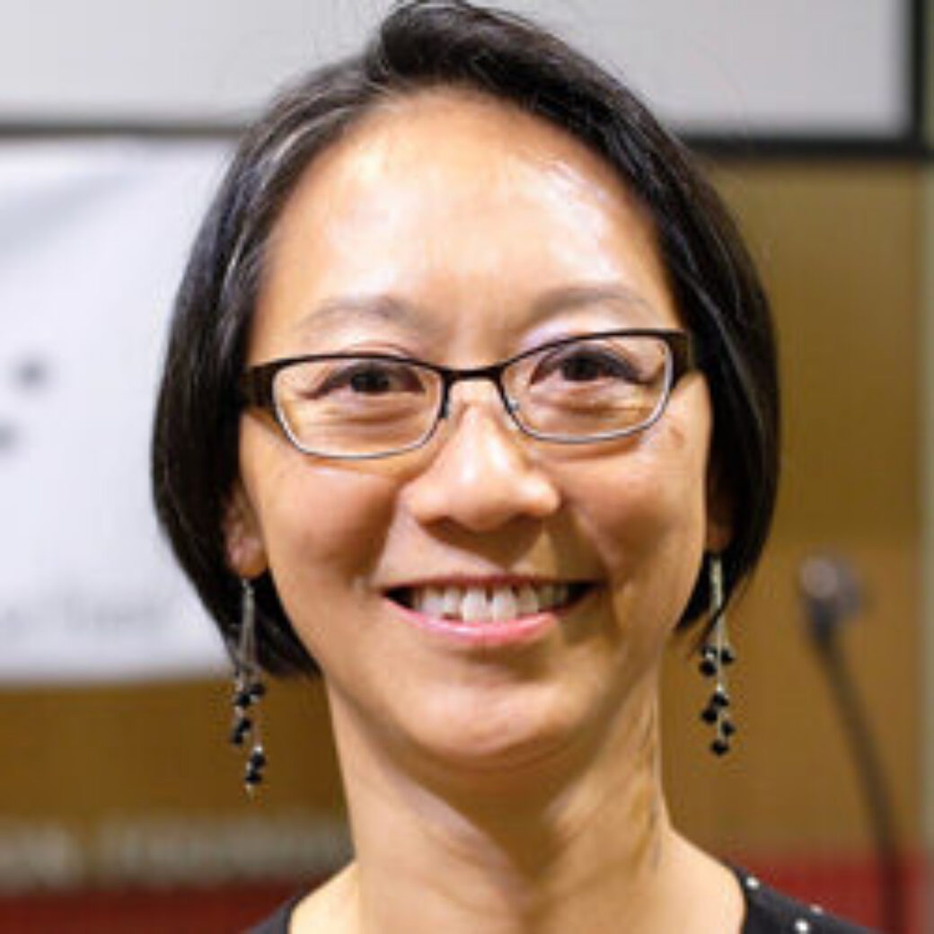 Close-up photo of Silvia, a middle-aged smiling Asian woman with chin-length black hair wearing glasses, a navy top with white polka dots, and dangling earrings.