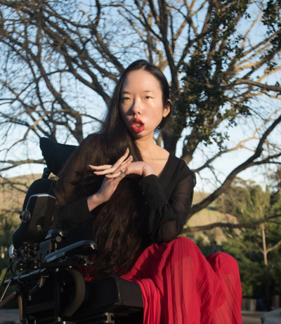 Aubrie Lee folds her hands and looks at the camera. She's riding a power wheelchair. Behind her, an oak tree.