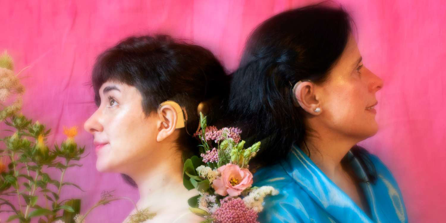Two people, heads back to back, with flowers in between them. One is a cochlear implant user, the other uses a hearing aid.