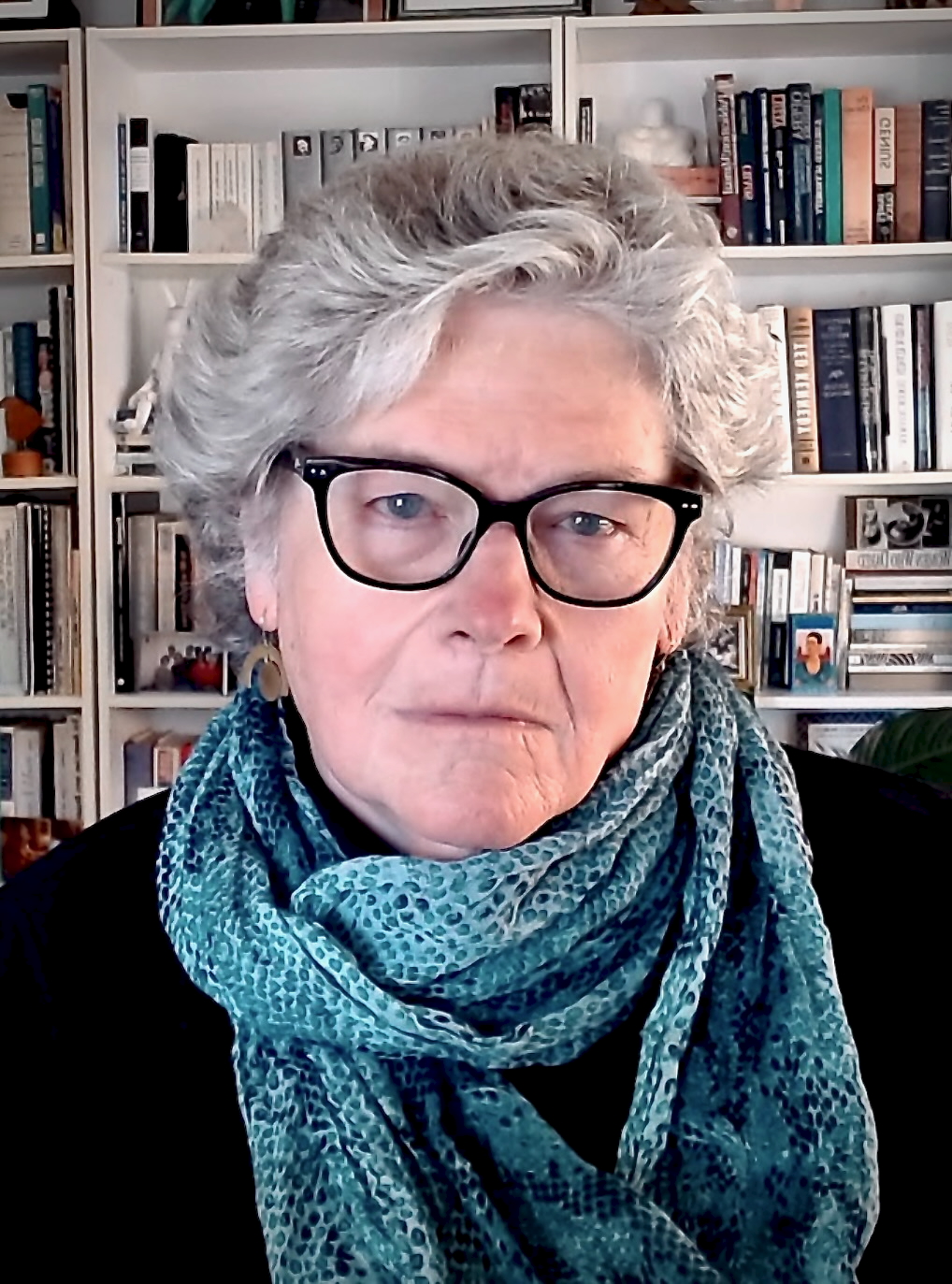 Portrait of Mary Lou, an older white woman with gray, wavy hair, wearing glasses, a blue-and-gray patterned scarf, and a black shirt. A bookshelf is in the background.