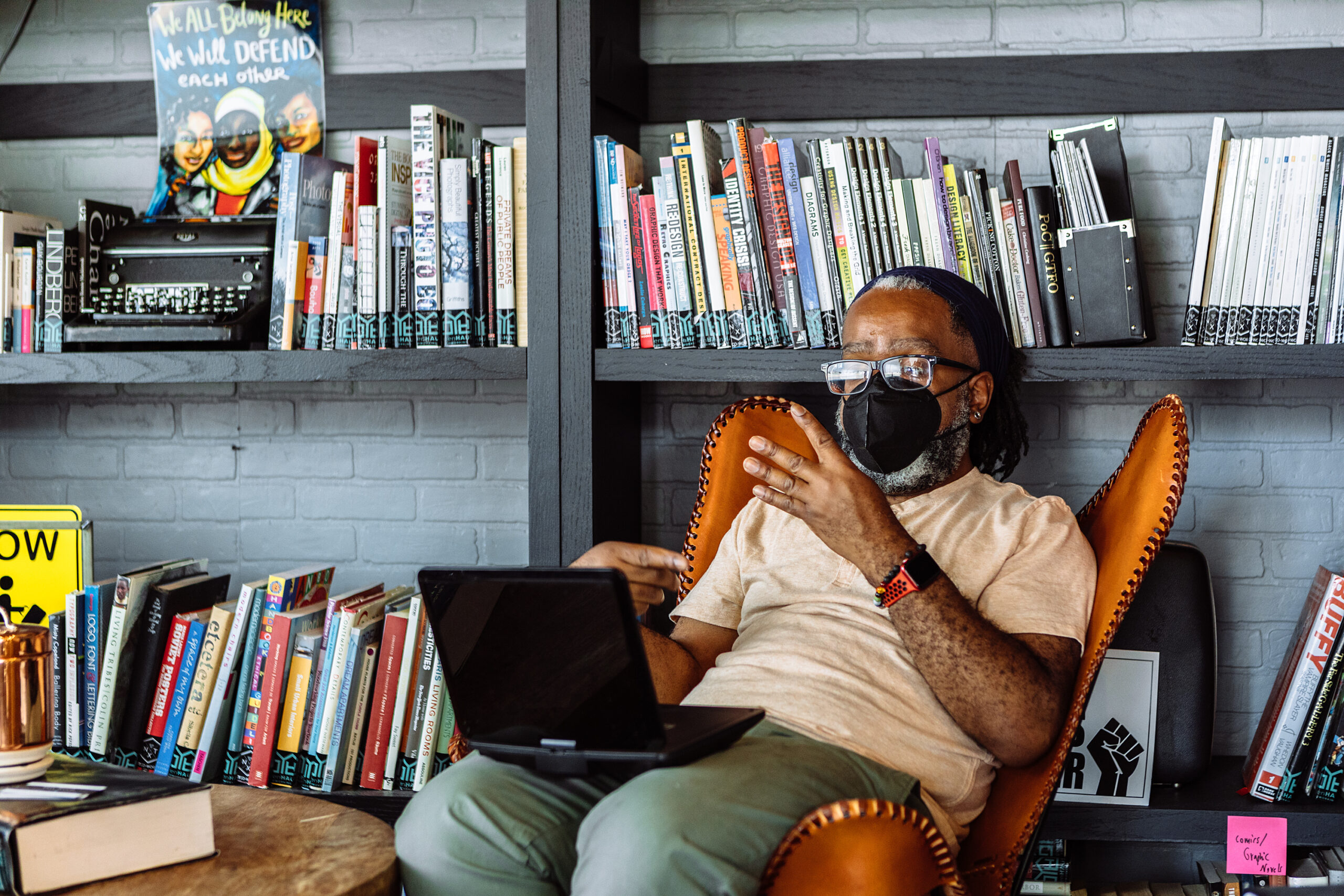 A Deaf Black man wearing glasses and a KN-95 mask leans back in a chair while signing in a video meeting via laptop. A community library filled with books and social justice posters serves as the backdrop.