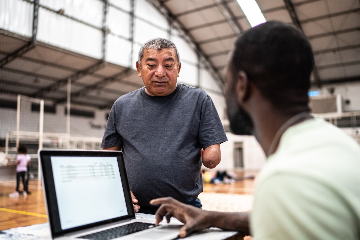 soldier at a laptop speaking with an older masculine hispanic presenting person in an emergency shelter