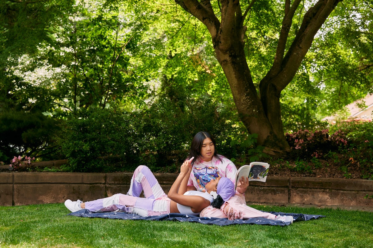 Wide shot of a young autistic couple practicing parallel play while lounging on a picnic blanket at a lush green park. On the left, the Black woman lies on her partner’s lap, concentrating on her popsocket fidget. On the right, the non-binary Asian person reads a poetry book.