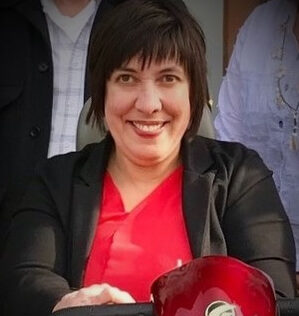 Portrait of Nicole Bohn, a white woman with short brown hair. She’s smiling directly at the camera in her red power scooter, wearing a red top with a black blazer.