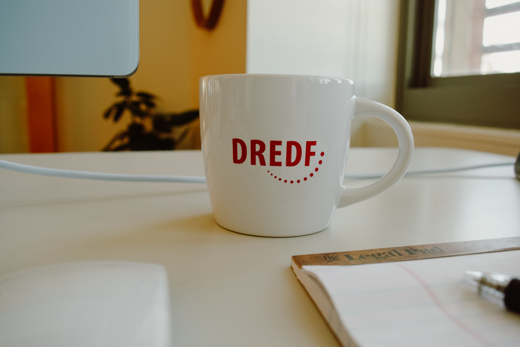 A white coffee mug that has the DREDF logo on it next to a legal pad