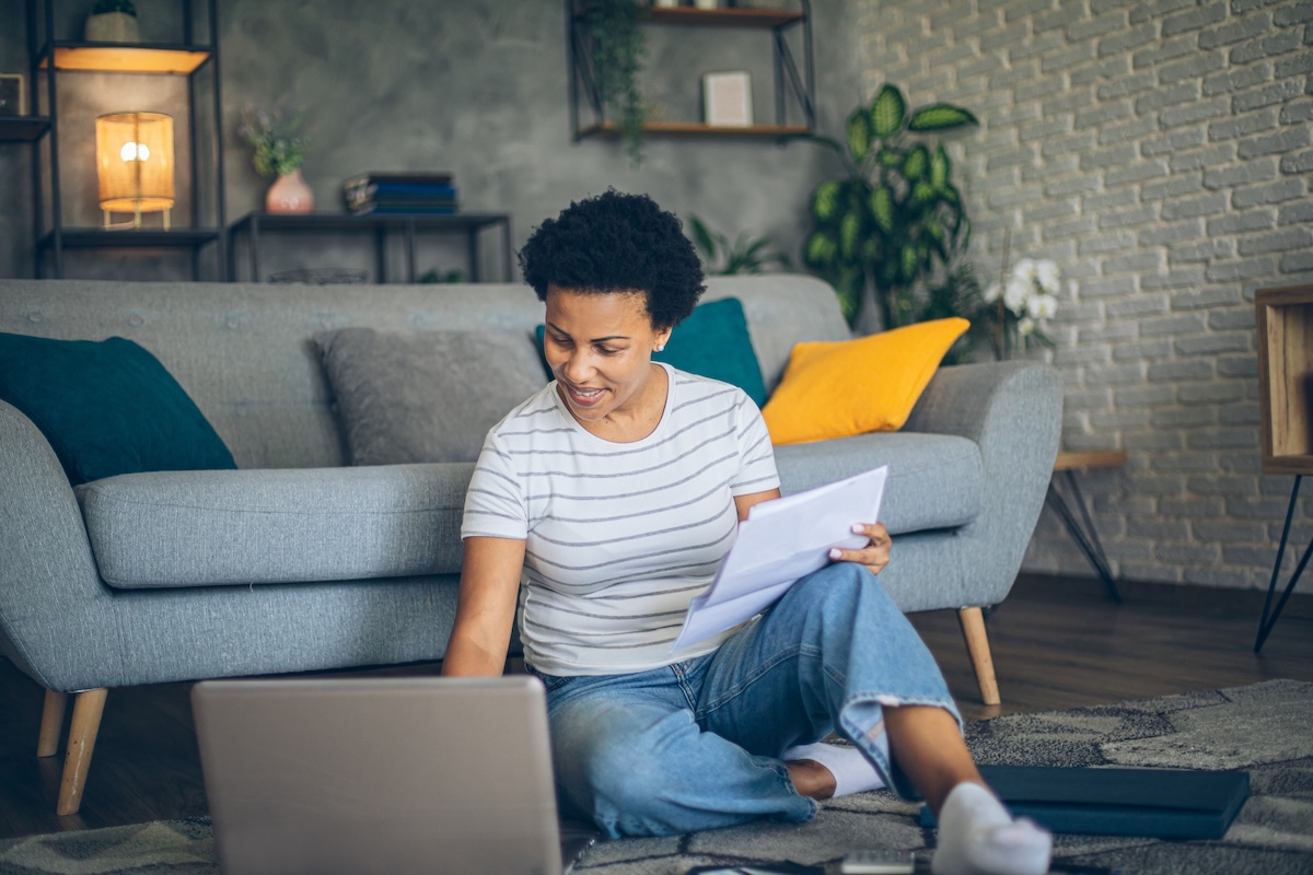 A female presenting person sits on a living room floor with a folder and a laptop