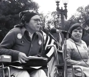 Kitty Cone speaking into a microphone at the demonstration. Judy Heumann is next to her on stage.