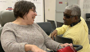 Nicole, a white woman in a red power scooter, and Dennis, a black man who is blind and wears sunglasses, smile at each other in conversation. A San Francisco Public Library backdrop is visible in the background. 