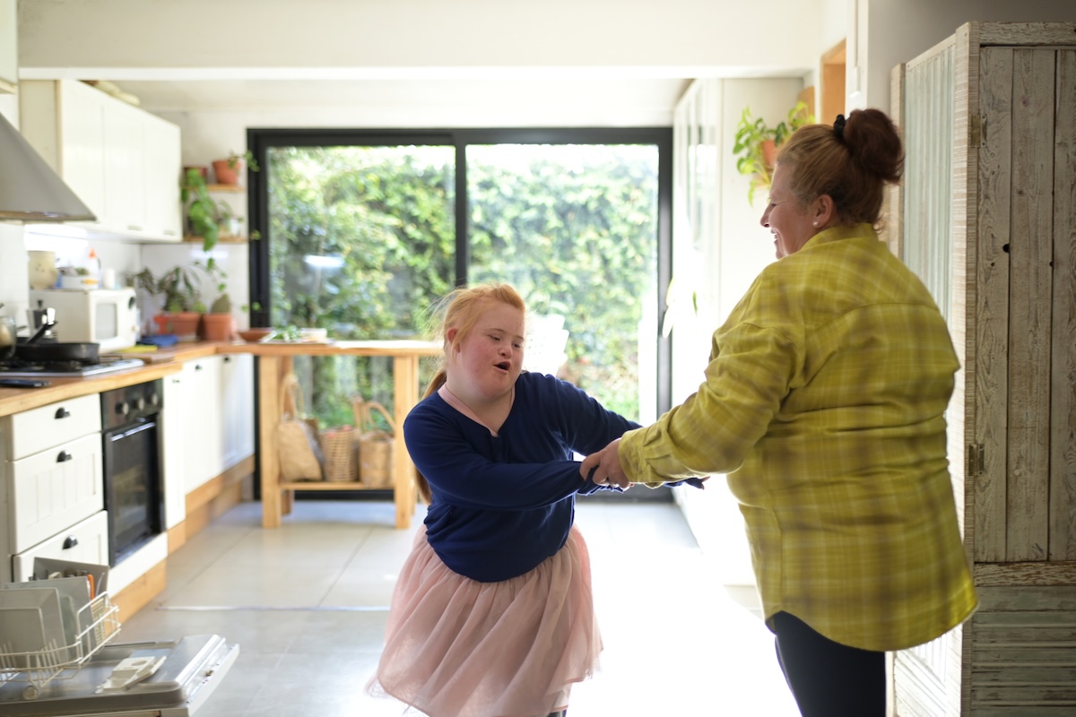 Young female presenting person with down syndrome dances with older family member in a bright kitchen.