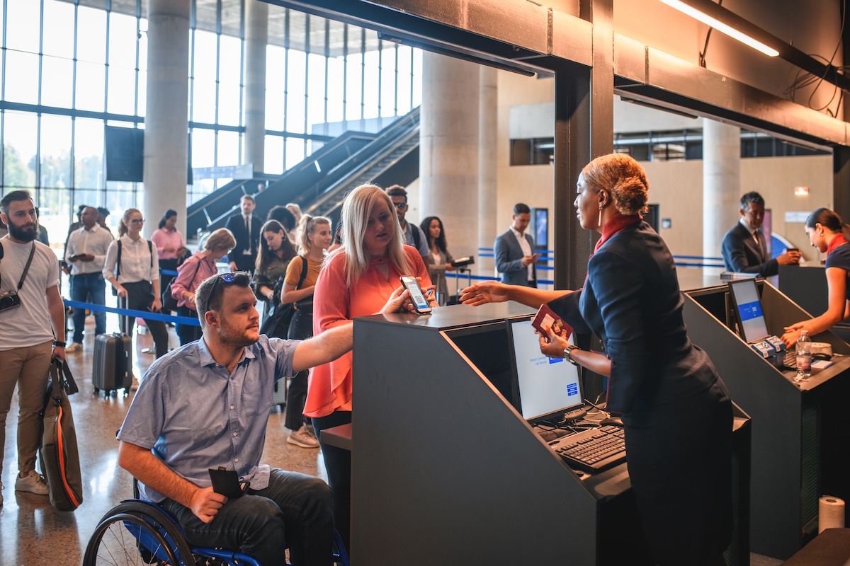 A masculine presenting wheelchair user presents papers to a airline worker at the airport