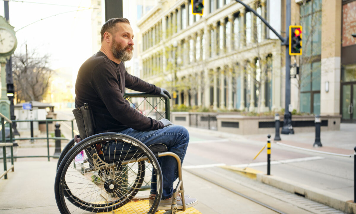 A white masculine presenting wheelchair user waits at a bus stop