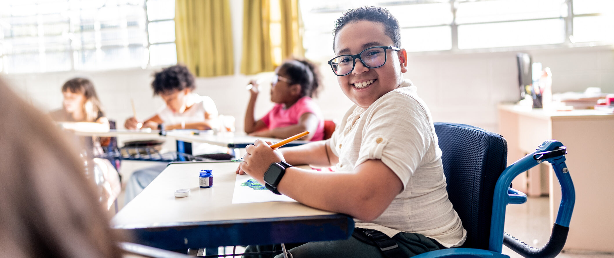 Young student that is a wheelchair user smiling to the camera in a sunny classroom