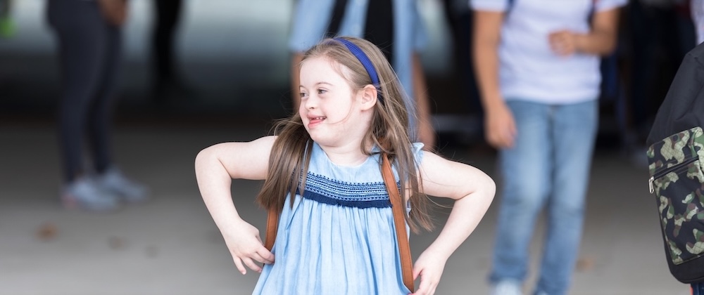 A young white girl with Down Syndrome in a backpack and a light blue dress.
