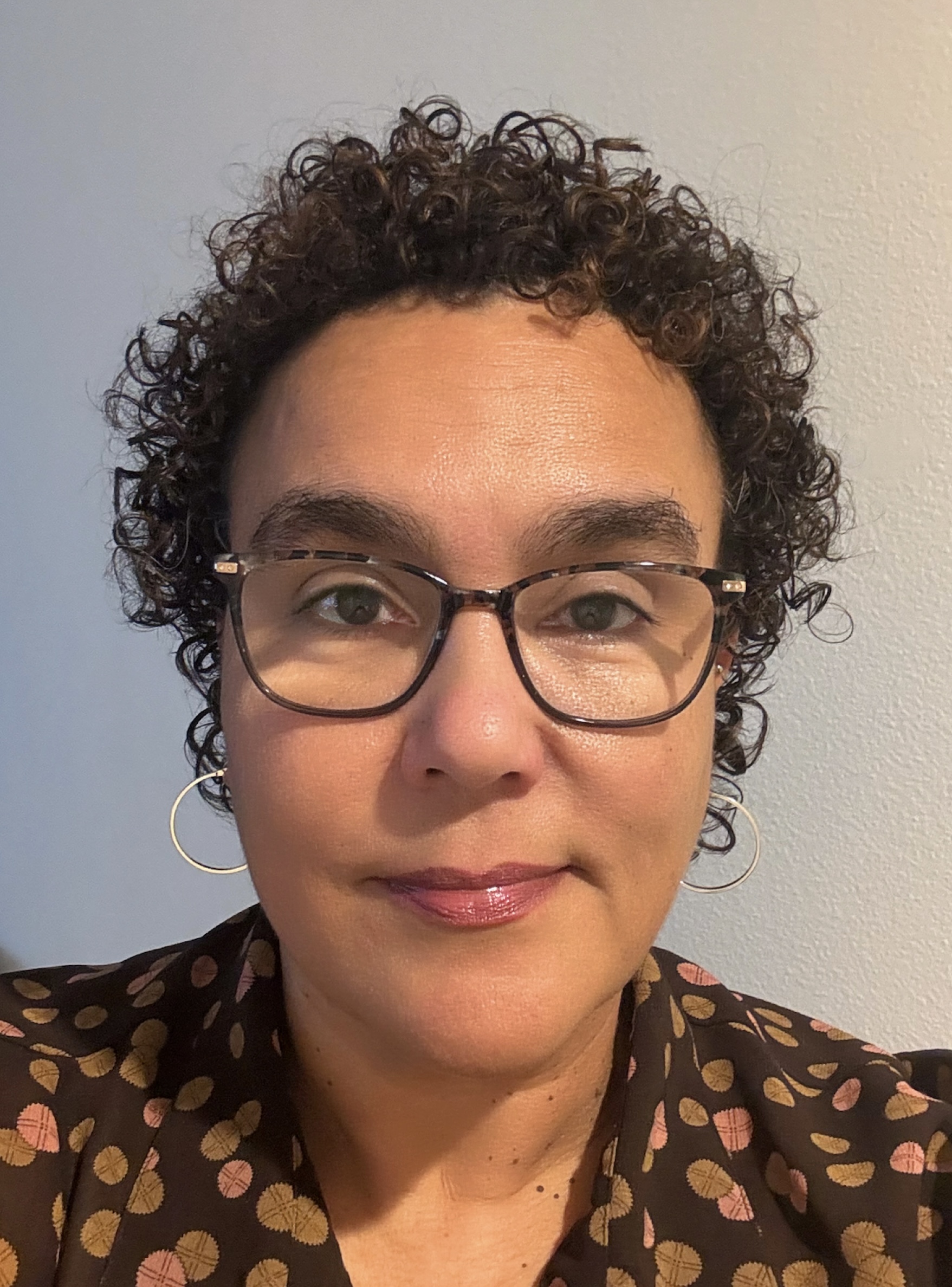 Portrait of Michelle Uzeta, a mixed race Black woman with curly brown hair. She smiles at the camera and wears glasses and a patterned top.