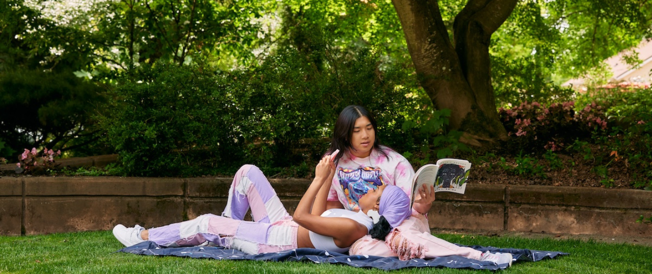 Wide shot of a young autistic couple practicing parallel play while lounging on a picnic blanket at a lush green park. On the left, the Black woman lies on her partner’s lap, concentrating on her popsocket fidget. On the right, the non-binary Asian person reads a poetry book.