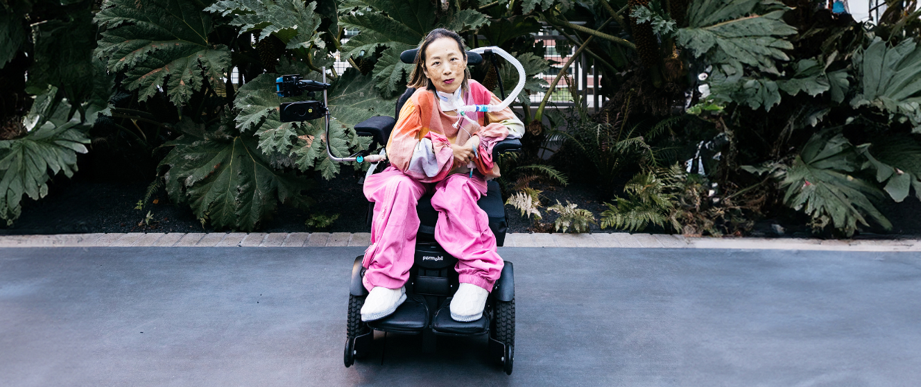 Asian American woman in a wheelchair with a tracheostomy at her neck that has a tube connected to her ventilator. Her head is tilted to the left side, she is smiling, and she is wearing a magenta lip color. Her shirt is plaid with pale pink and other pastel colors and she is wearing pink pants. Behind her is a lot of greenery from bushes and trees at a park. A dark color fades over the photo. Photo Credit: John D. and Catherine T. MacArthur Foundation.