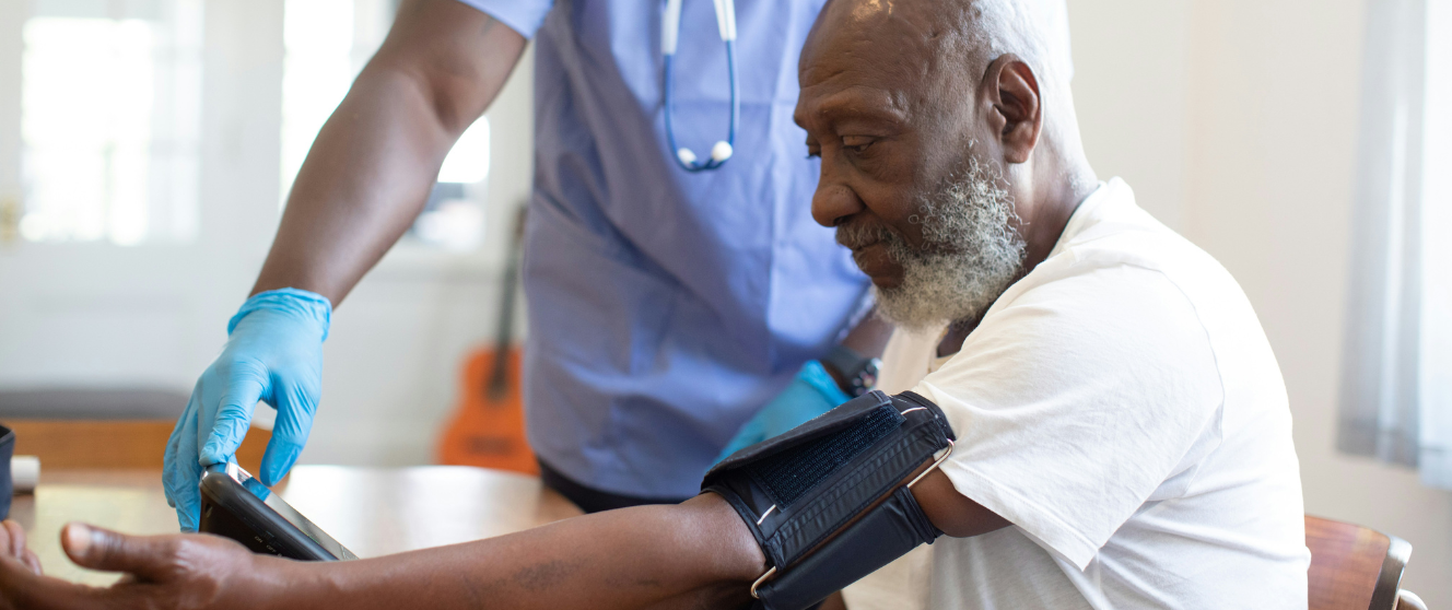 A Black, masculine presenting person, gets their blood pressure read by a medical provider in blue scrubs