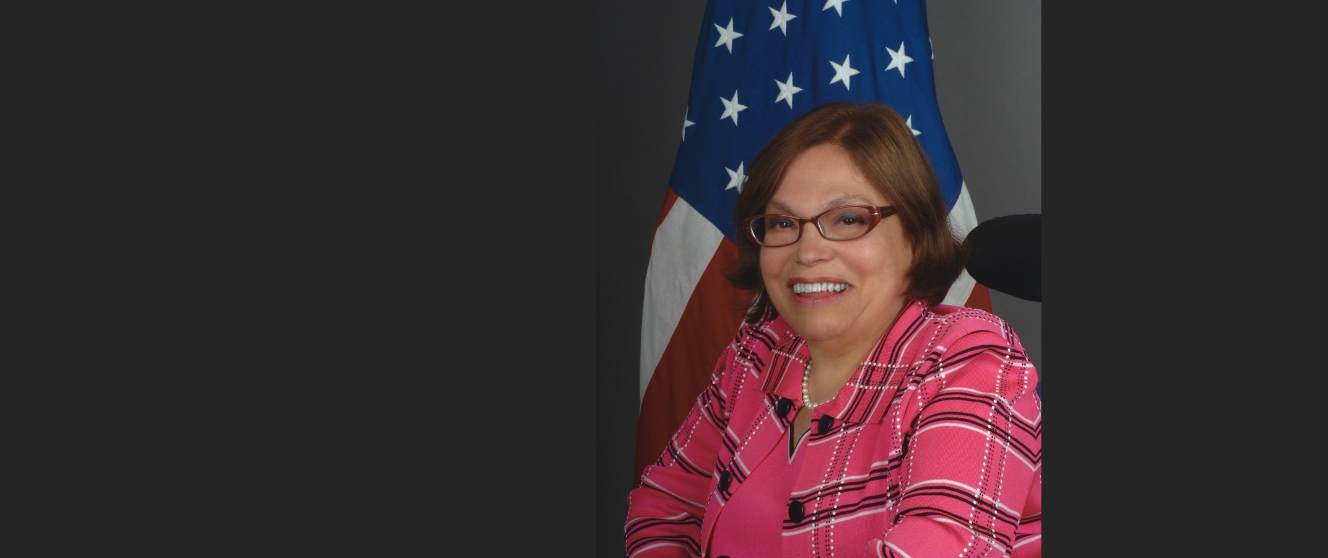 Portrait of Judy Heumann, an older white woman in a pink plaid top in front of the American flag