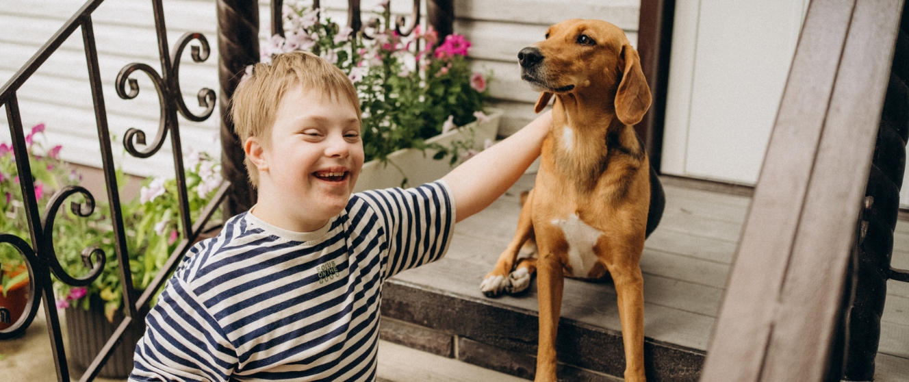 A young white masculine presenting person with Down Syndrome sits on outdoor steps to a home petting a dog.