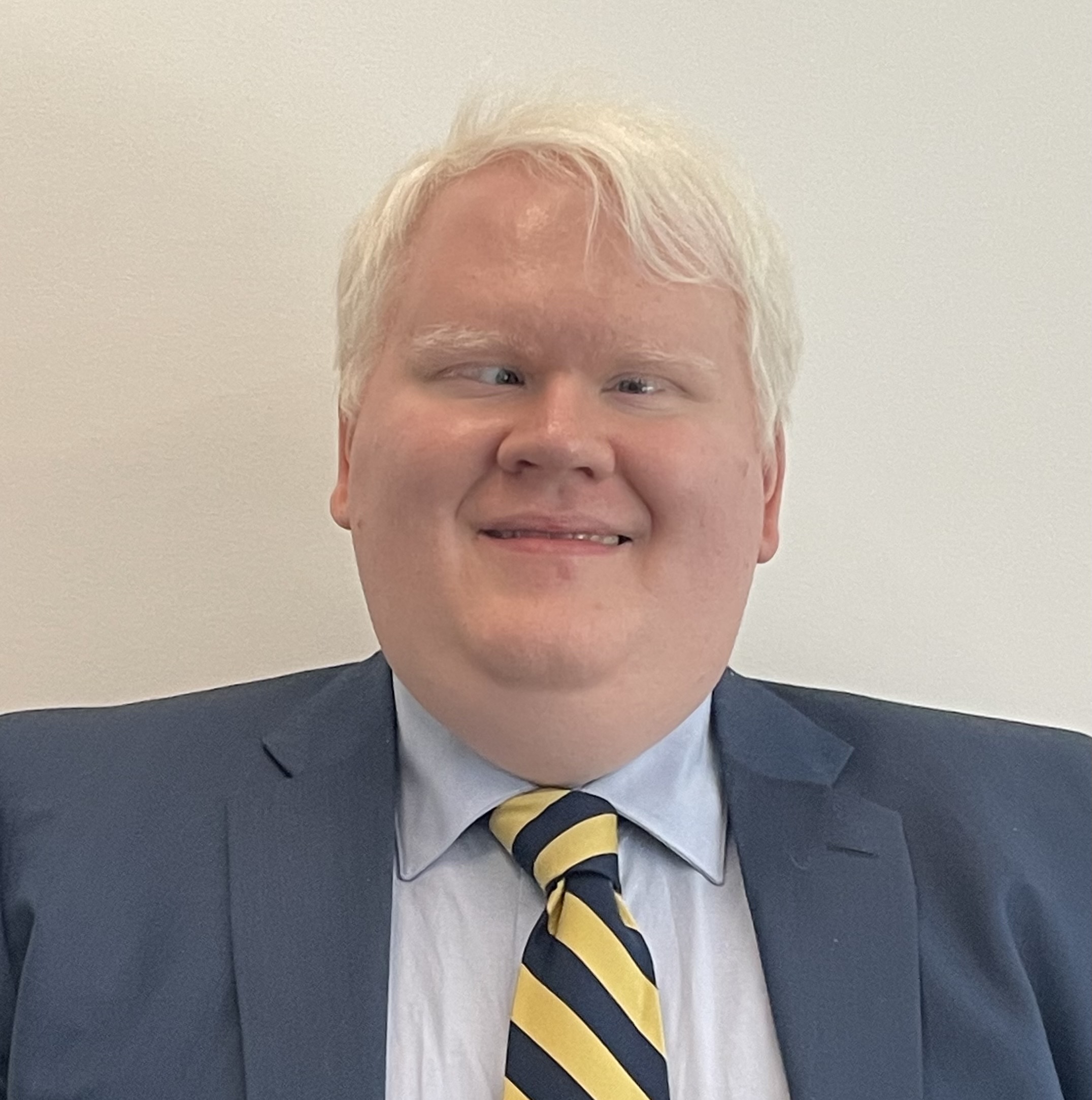 A portrait of Brian, a man with Albinism in his 30s wearing a blue suit and maize and blue striped tie.