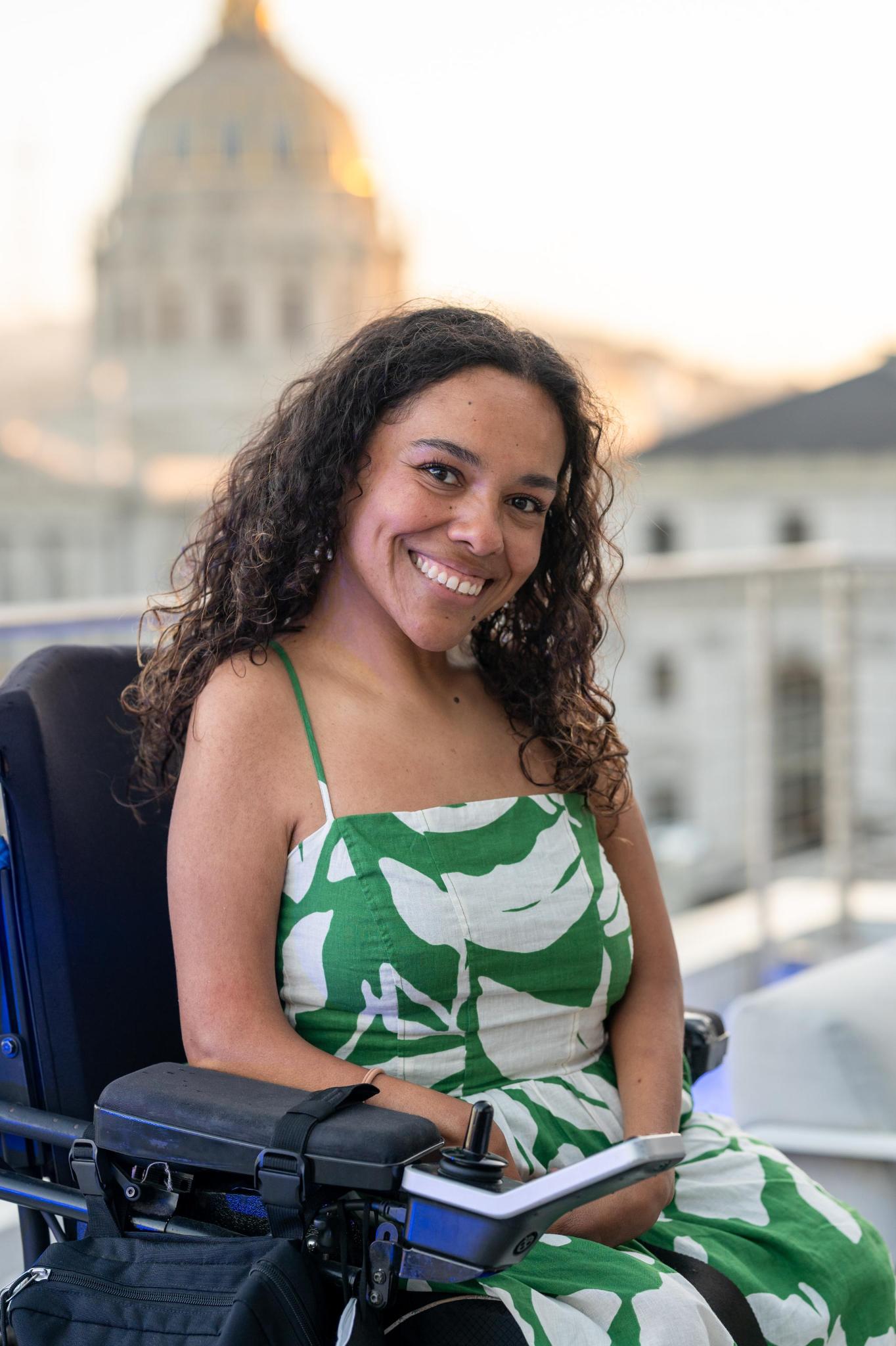 Brittanie, has brown skin and long, dark curly hair, sits in her power wheelchair wearing a green-and-white patterned dress. The San Francisco skyline is softly blurred in the background.