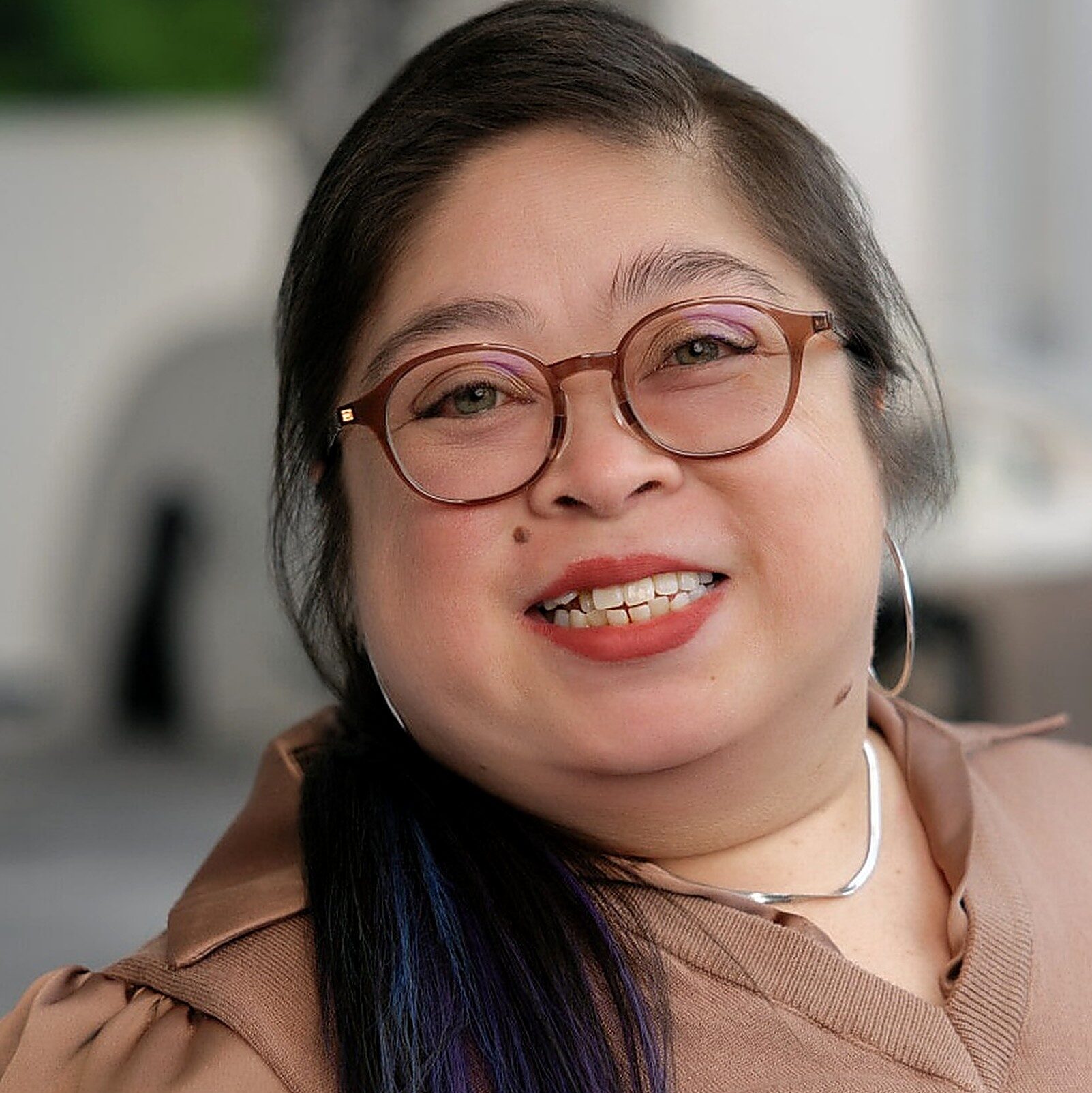 A portrait of a dark-haired woman in her forties with blue-purple ombré tips, round amber glasses, smiling, coral lipstick, tan top, blurred office background.