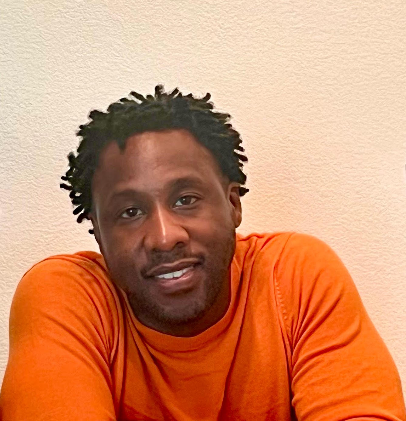 A Potrait of Nel in an orange sweater, black man his early 40s with short dreadlocs.