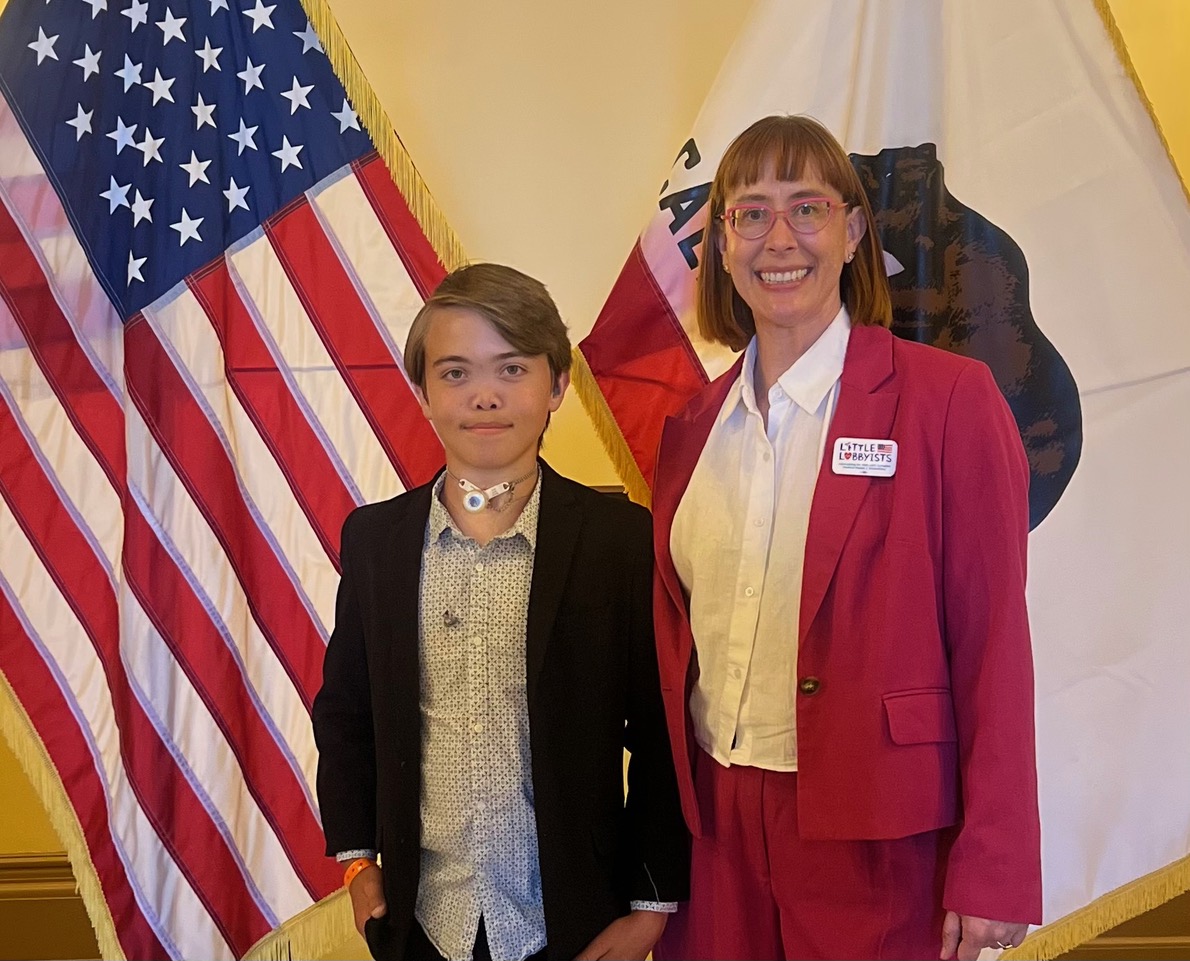 A portrait of two people standing in front of an American and California flag. On the left is James, a white teen boy wearing a black suit. On the right is Jenny, a white woman in her forties with pink glasses, bangs, and a bob haircut wearing a pink suit.