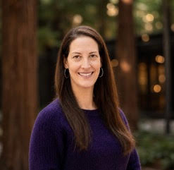 A portrait of Rachel, a white woman with long brown hair, smiling and wearing a purple sweater, photographed outdoors among trees.