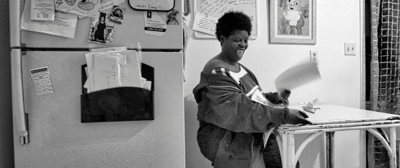 Lois Curtis, plaintiff in Olmstead v. Curtis, smiles while holding papers in a home setting, with a refrigerator, notes, artwork, and other everyday household items behind her.