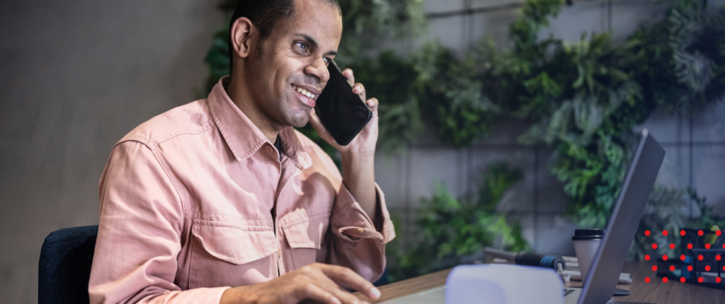 A masculine presenting person smiles on a laptop while on a smart phone