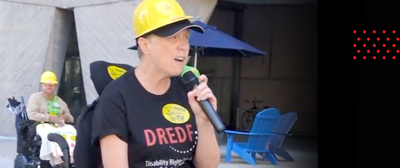 CT wearing a DREDF tshirt and a yellow hard hat with a microphone in their hand